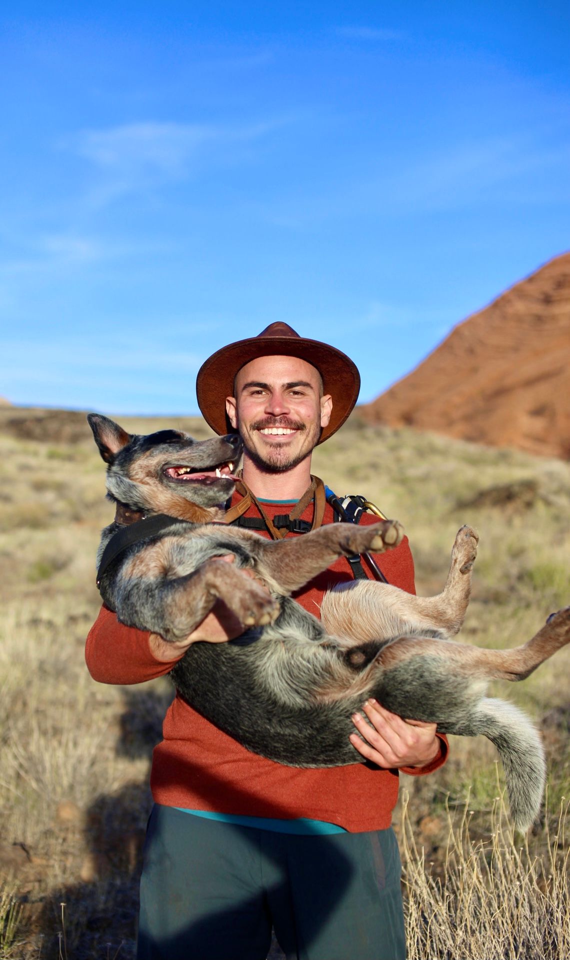 Nick Elisseos in a wide-brimmed hat, smiling and holding his blue heeler in both arms, on open grassland under a clear sky.